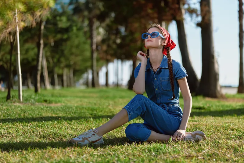 young woman sitting on grass wearing blue jumpsuit and red headscarf outdoors in sunny park with row of trees and green lawn