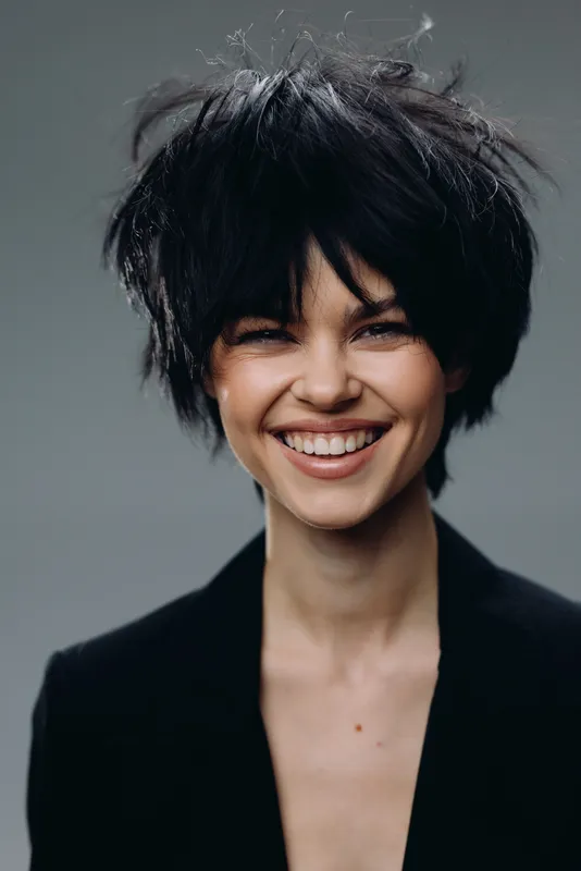 Smiling woman with messy black hair wearing a black jacket, expressing happiness and confidence in a studio setting.