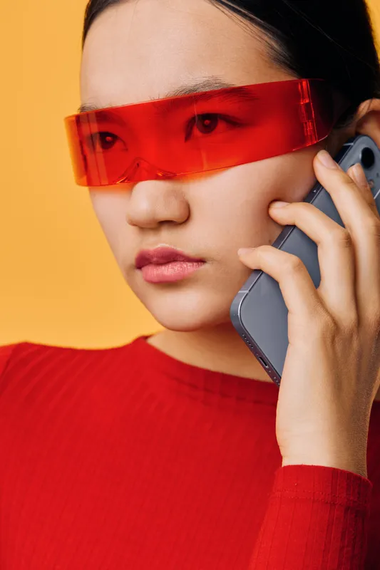 Young woman with red futuristic glasses talking on smartphone against orange background. Closeup of female in red sweater with modern eyewear in communication and technology concept.