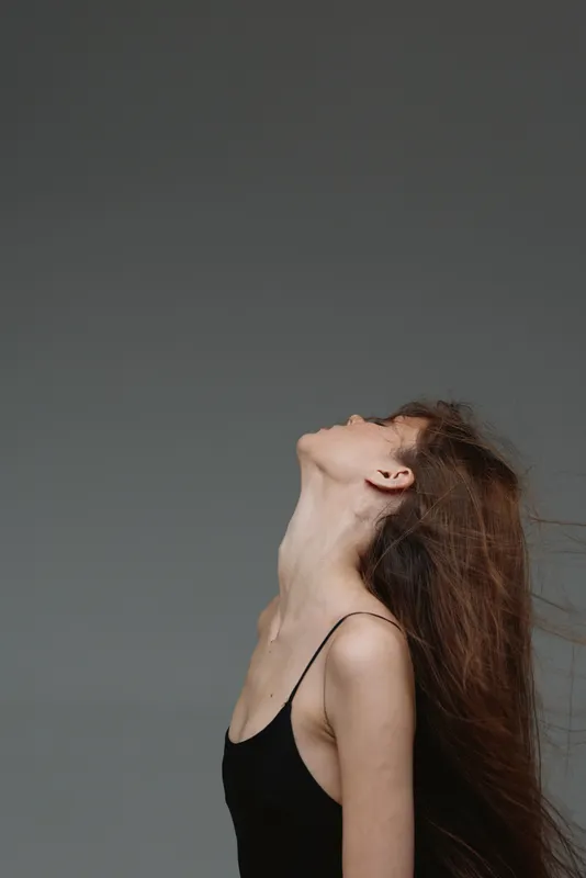 Young woman with long hair wearing black top looking up on gray background with minimalist style.