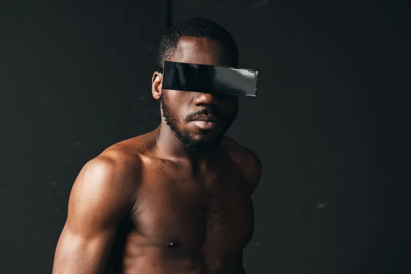African man shirtless with muscular body and black tape covering eyes in creative conceptual visual style on dark background for artistic expression and mystery.