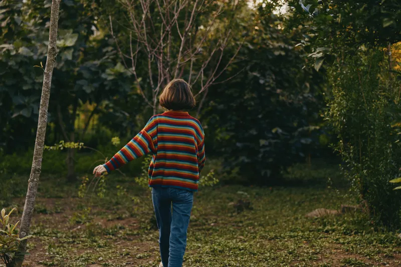 Child 9-year-old with short brown hair in a colorful striped sweater walking on a forest path, casual outfit and jeans, back view exploring peaceful nature and greenery.