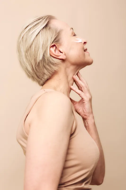 Profile of mature woman with short blonde hair applying face cream on cheek, touching neck gently. Concept of skincare, aging skin care, beauty routine, wellness and self-care.