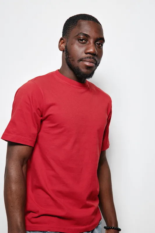 African American man with dark skin wearing red t shirt standing against white background looking at camera with confident expression and casual posture.