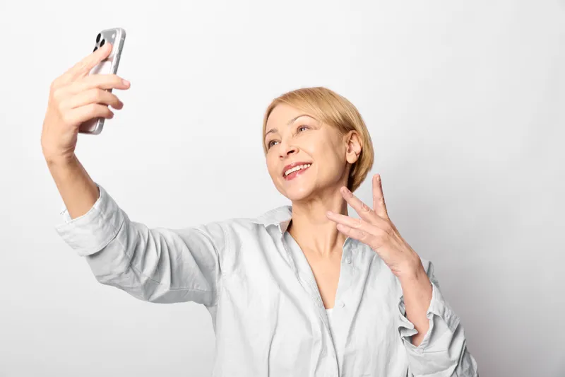 Smiling woman takes a selfie with a smartphone,peace gesture,studio portrait on white background,casual outfit,showing friendly communication and joy
