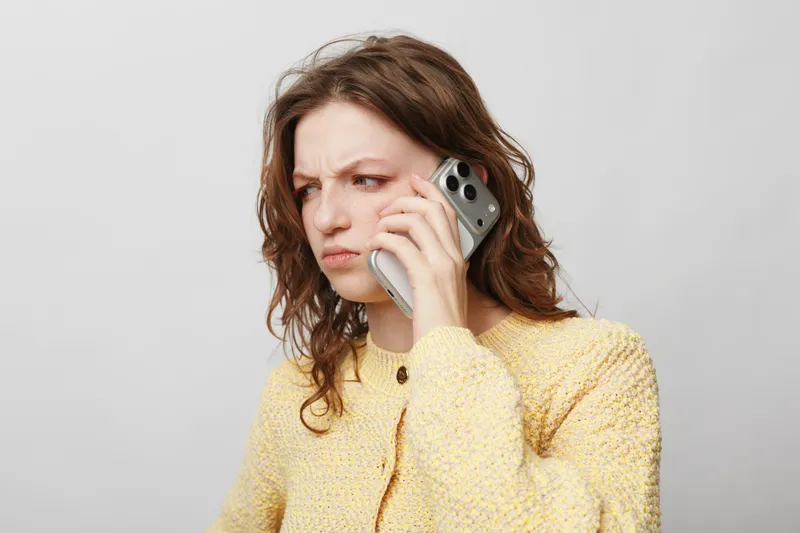 Concerned woman with curly hair wearing yellow sweater talks on smartphone. She looks worried and focused against plain light background. Concept of communication, stress, and emotion.