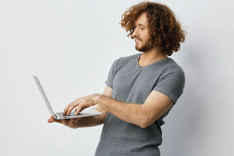 Young man with curly hair using a laptop, wearing a grey shirt, smiling in a minimalistic indoor setting, perfect for concepts like remote work and technology