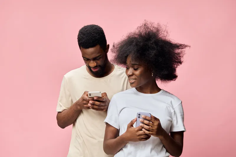 Young Black couple engaged with smartphones, showcasing casual fashion and joyful expressions against a soft pink background