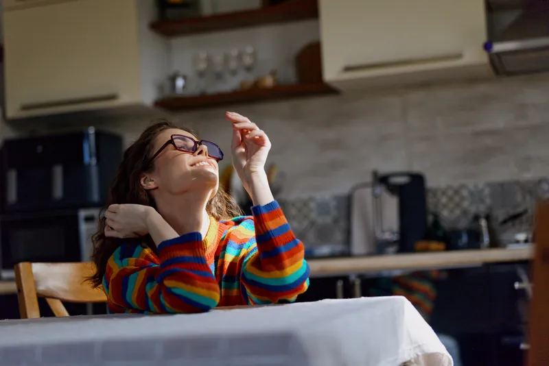 A thoughtful woman sits at a dining table in a colorful striped sweater, glasses raised toward the ceiling, reflecting on ideas in a bright home kitchen setting.