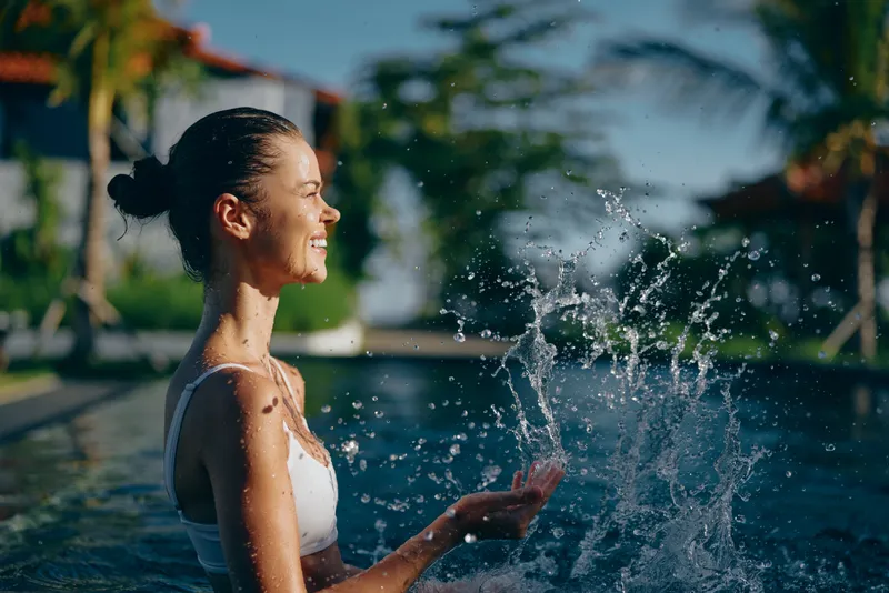 Cheerful woman in a white swimsuit stands in a sunlit pool, water splashing as she laughs, tropical garden backdrop, relaxed posture, carefree outdoor lifestyle amid lush greenery and blue skies