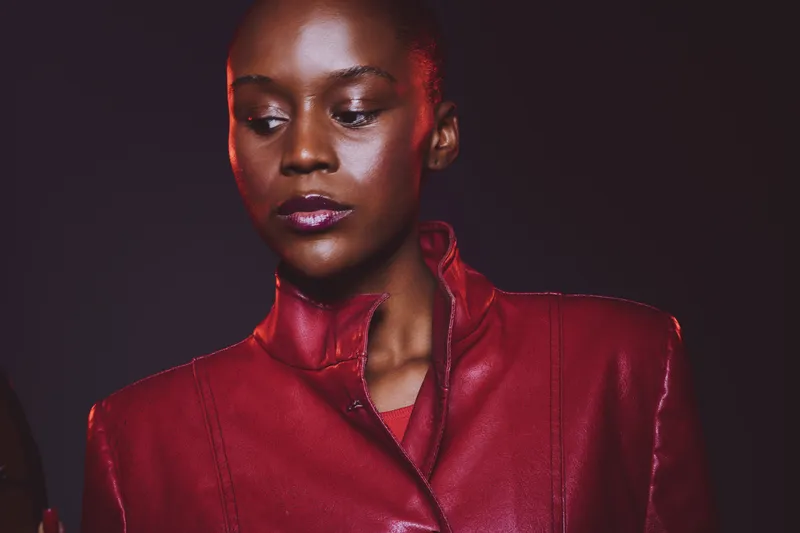 Portrait of a confident dark skinned African woman wearing a red leather jacket, looking down with a calm expression. Studio lighting highlights her smooth skin and elegant style.