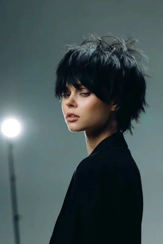 young woman with short black hair posing indoors wearing dark jacket with moody lighting