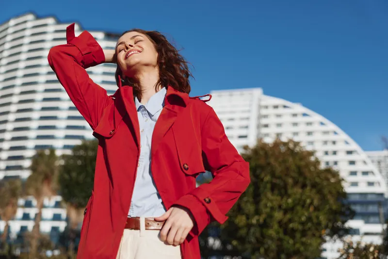 Young woman in a red coat enjoys the sunny day outdoors with her eyes closed and a joyful smile on her face against a modern urban background.