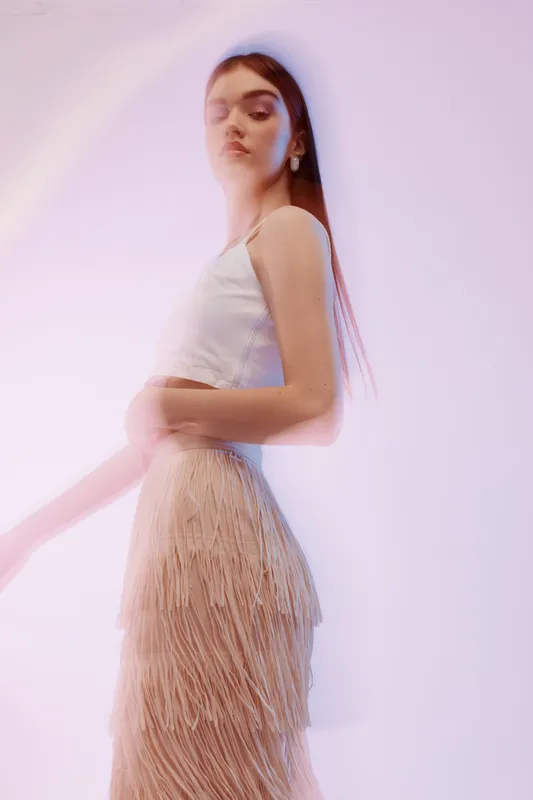 Young woman in a white top and fringed skirt poses with closed eyes in soft pink studio lighting. Concept for beauty, fashion, elegance, and modern style.