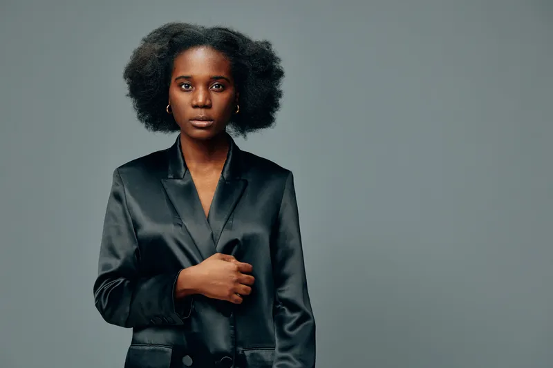 Confident model with natural hair in a black satin blazer against a minimalist grey background, exuding elegance and strength