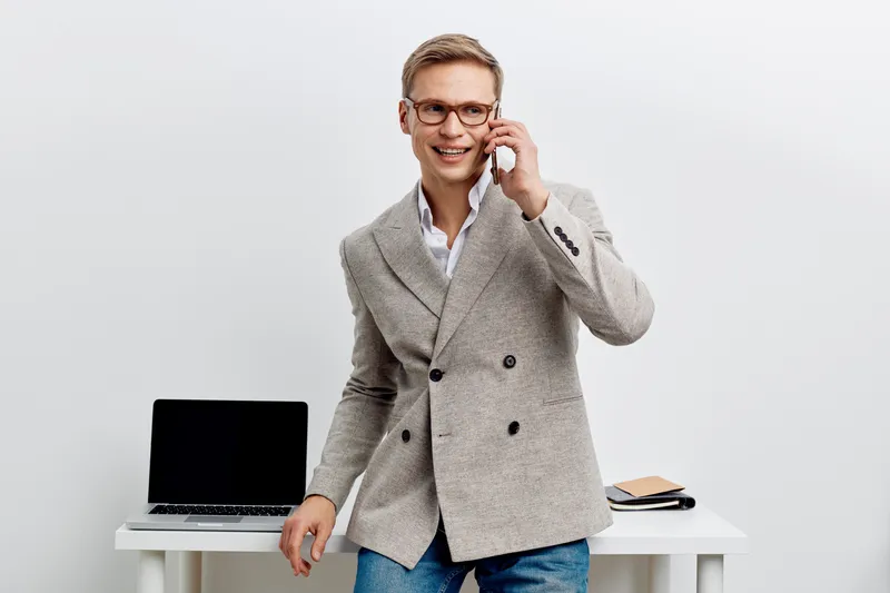 Professional young Caucasian man in smart casual blazer and glasses talking on mobile phone, standing in modern office with laptop and documents, smiling, cheerful, workplace scene, business casual st