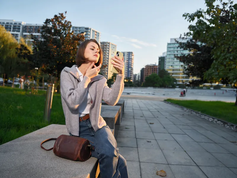 A cheerful woman sits on a concrete bench in a modern urban park, texting on her phone. She smiles warmly, appears confident, and relaxes in bright daylight within a vibrant city setting.