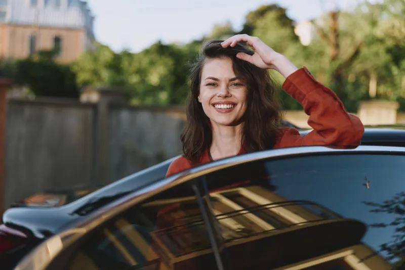 A smiling woman leans on a sleek car in a sunny outdoor setting, showcasing casual lifestyle and confident expression. A bright street scene of everyday joy today