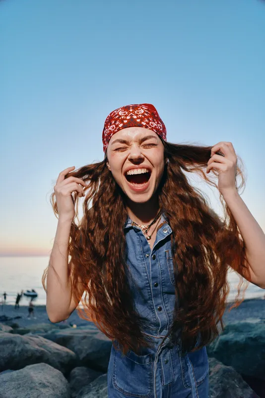 Lifestyle woman with long curly hair wearing modern boho-western denim street style and red bandana, laughing joyfully at the beach during golden hour, film color effect.