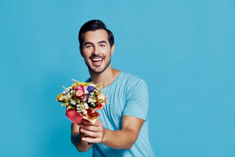 Joyful smiling man holding a colorful bouquet of flowers, standing against a bright blue background. Cheerful expression reflecting happiness and positivity.