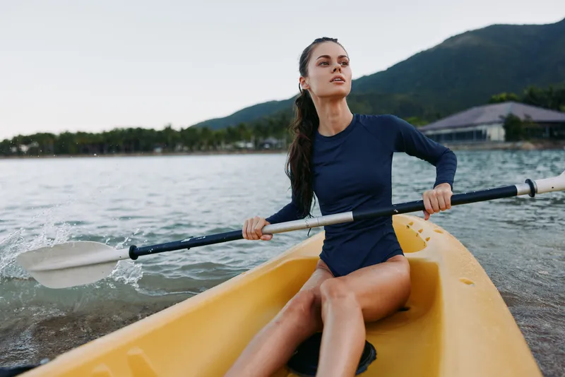 A lone female kayaker sits in a bright yellow kayak, paddling across calm water in a navy long sleeve top, with a focused gaze and a tranquil lakeside backdrop.