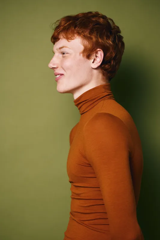 Profile of young man with red hair wearing orange turtleneck sweater standing against green background looking ahead with slight smile expression in studio portrait shot.
