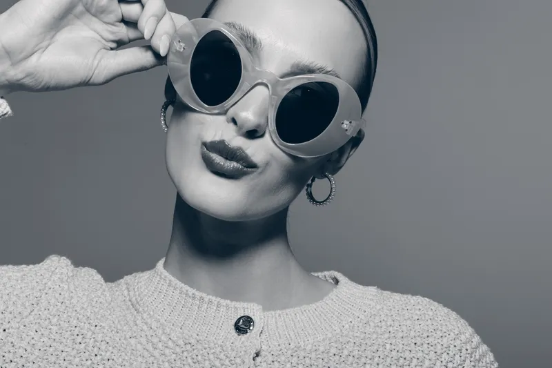 Stylish young white woman with pale skin wears round sunglasses and hoop earrings, posing with puckered lips in monochrome studio setting. Concept of fashion, attitude, and accessories.