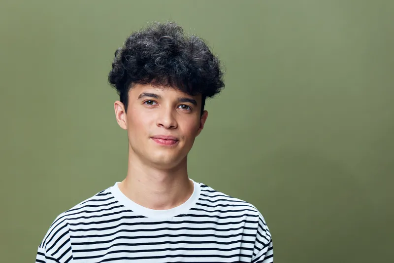 Young man with curly hair wearing striped shirt standing against green background, looking confident and approachable in casual style portrait.