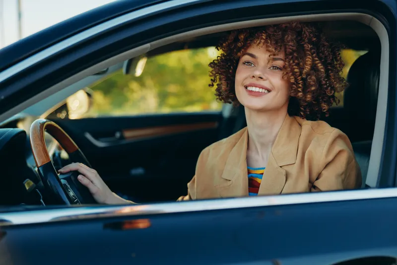 Cheerful woman driver in a car, smiling inside the sunlit interior, retro film color mood, lifestyle moment, relaxed posture, natural tones, candid joy