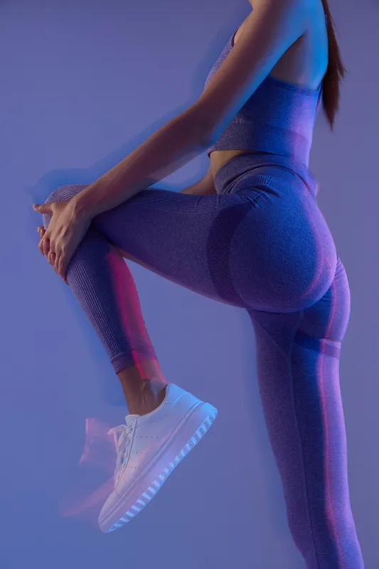 woman sport fitness in purple activewear stretching her leg with white sneakers under vibrant blue and pink lighting, emphasizing strength and flexibility