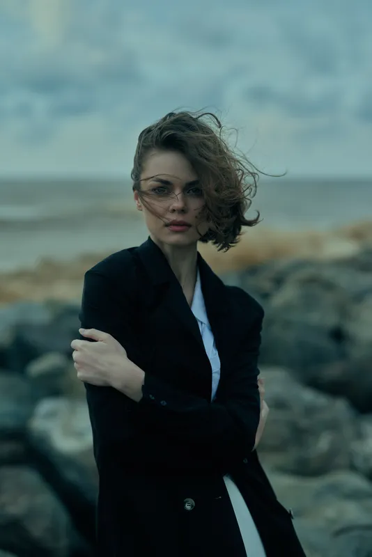 Woman in a blazer stands on a windy seaside, arms crossed, serious expression, rocky shore and moody sky, fashion mood for outdoor editorial use.