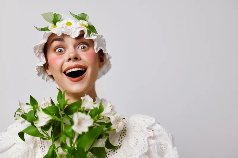 Cheerful young woman wearing a flower crown and white dress, holding a bouquet of flowers against a light background, exuding joy and positivity