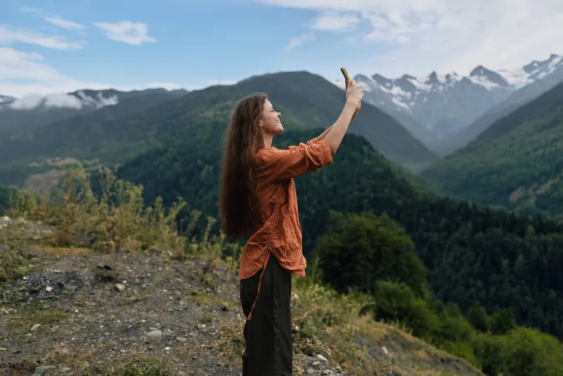 Woman stands on mountain overlook using smartphone to photograph landscape nature and mountainview during hiking outdoors trip, authentic inclusive portrait of adventure and travel