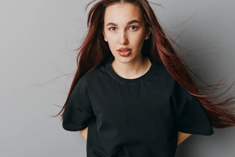 Young woman with long brown hair wearing black t shirt standing against gray background looking at camera with confident expression and natural makeup in studio portrait.