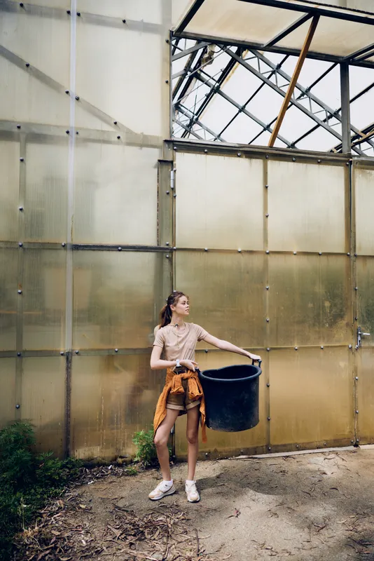 Woman holds a bucket in a greenhouse while gardening, casual young adult wearing sneakers and sweater tied at waist, standing near translucent panels with dirt and plants.