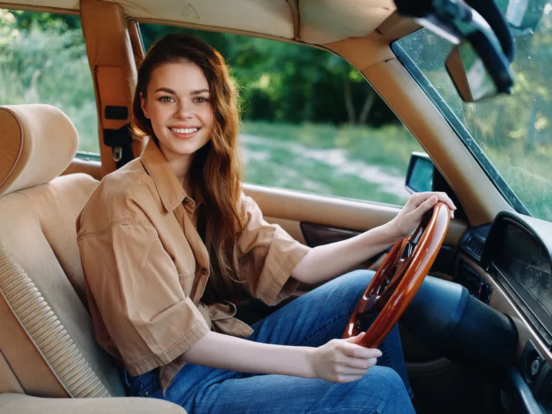 Young woman smiling while driving a vintage car in nature, wearing a casual brown shirt and jeans, surrounded by greenery and sunlight.