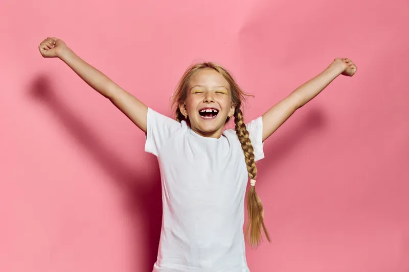 Happy girl with long braid wearing white t-shirt smiling and raising arms against pink background. Studio shot with bright lighting. Concept of joy, childhood, and freedom.