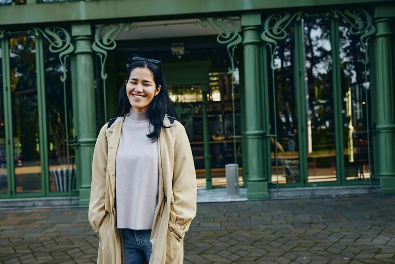 Woman wearing a beige coat and light sweater stands outdoors in front of a green cafe facade, smiling, casual street style urban backdrop for fashion photography