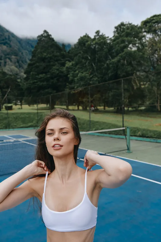 Young woman in a white sports bra on a tennis court, demonstrating confidence and fitness against a lush green background with mountains, ideal for healthy lifestyle themes