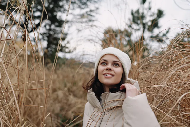 Woman in a warm beige coat and knit hat stands among tall dried grasses, outdoor winter scene, soft natural light, serene mood and subtle fashion sense.