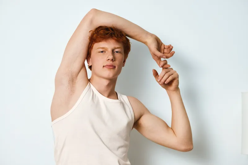 Young man with red hair and fair skin posing in white tank top stretching arms against white wall in minimal indoor setting. Confident posture and casual look for lifestyle portrait.