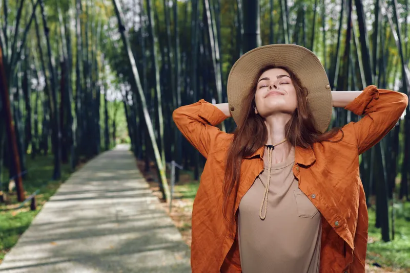 Woman in hat relax on forest path, eyes closed and arms behind head, wearing orange coat and knit sweater. Nature escape, calm moment, outdoor leisure and fresh air.