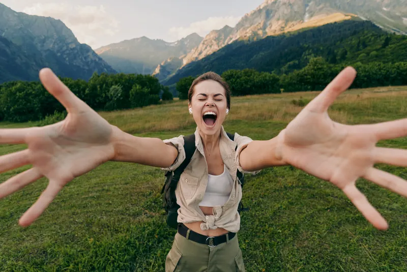 Smiling woman with outstretched arms in a lush valley, mountains in the background. Lifestyle outdoors vibe, happiness, nature, adventure, and travel inspiration.