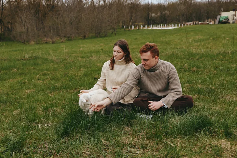 Young couple in warm sweaters sits on grass in an open field petting a white baby goat enjoying a calm and natural lifestyle moment in early spring outdoors.