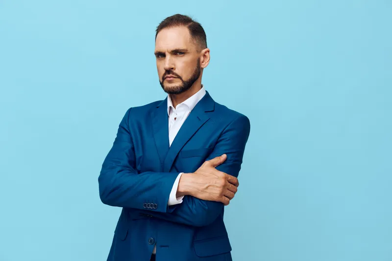 A professional man stands against an isolated blue background, exuding confidence and focus. He wears a tailored suit, arms crossed, with a calm, assertive demeanor.