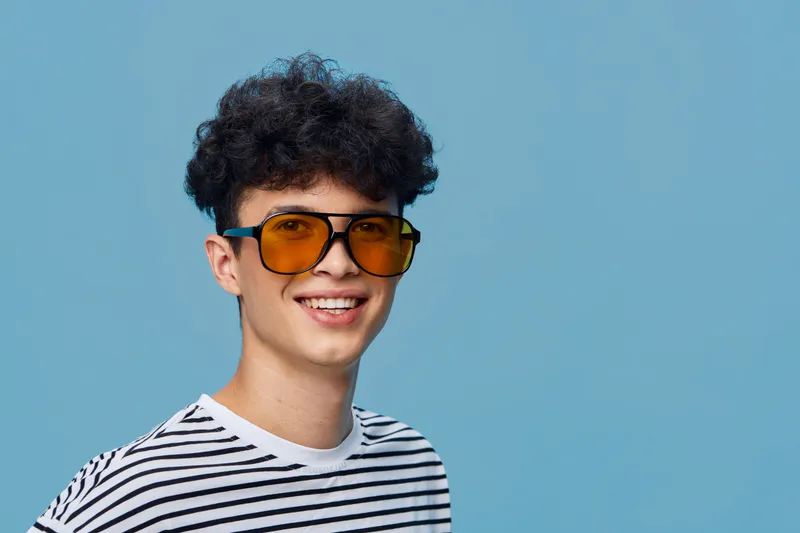 smiling man wearing yellow sunglasses and a striped shirt posing against blue background, casual style with short curly hair and cheerful expression