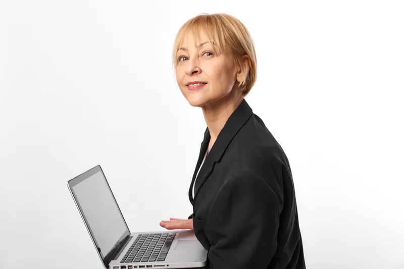 Mature woman in business blazer works at laptop in office portrait, facing camera, calm professional technology communication concept for remote work productivity