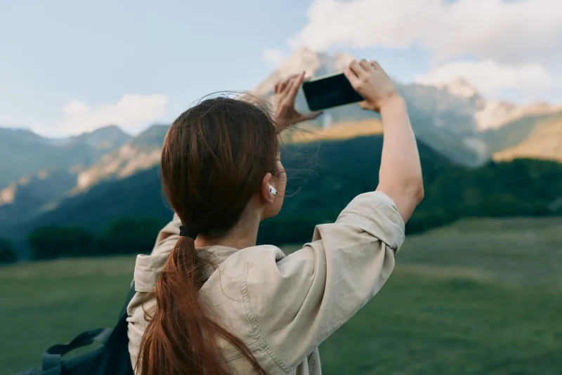 Woman outdoors taking a photo with a smartphone, backpacked casual style, lush green field and distant mountains, bright natural light, relaxed lifestyle moment under clear blue sky