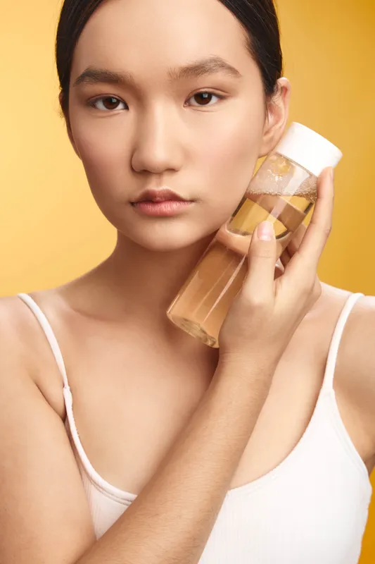 asian woman holds a clear bottle near her face, skincare product in focus, soft lighting, calm expression, clean setting, beauty routine mood and serene atmosphere.