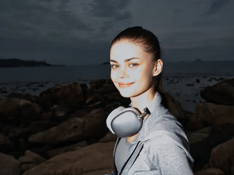 Young woman in sporty attire with headphones, smiling near rocky beach, showcasing a relaxed lifestyle and connection to nature, ideal for wellness concepts